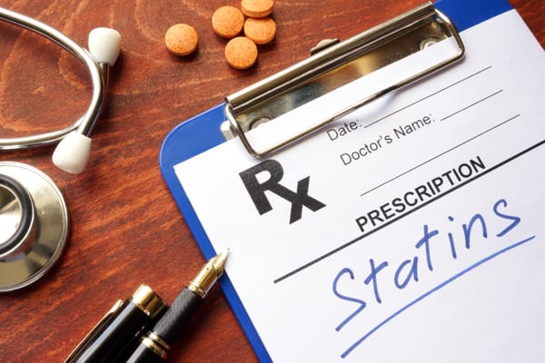 "Statins" written on prescription pad. Pens, pills, and stethoscope in background. 